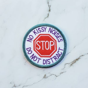 May include: A round, teal-colored embroidered patch with a white background. The patch features a red stop sign with the word "STOP" in white text. The text around the stop sign reads "No Kissy Noises Do Not Distract" in purple text.