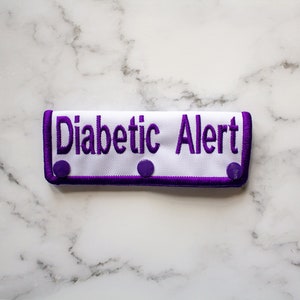 May include: White fabric patch with purple trim and the text "Diabetic Alert" in purple embroidery. The patch has three purple snap buttons.