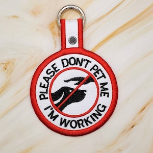 May include: Red and white round embroidered patch with a black silhouette of a dog with a red line through it. The text on the patch reads "Please Don't Pet Me I'm Working".