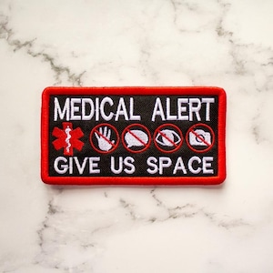 May include: Red and black embroidered patch with the text "MEDICAL ALERT GIVE US SPACE". The patch features a medical symbol, a hand with a slash through it, a speech bubble with a slash through it, an eye with a slash through it, and a camera with a slash through it.