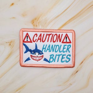 May include: A white embroidered patch with a pink border. The patch features a cartoon shark with a toothy grin and the text "CAUTION HANDLER BITES" in red and blue lettering.