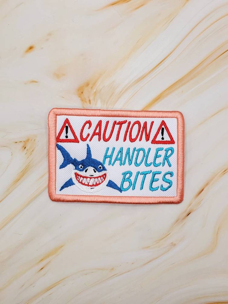 May include: A white embroidered patch with a pink border. The patch features a cartoon shark with a toothy grin and the text "CAUTION HANDLER BITES" in red and blue lettering.