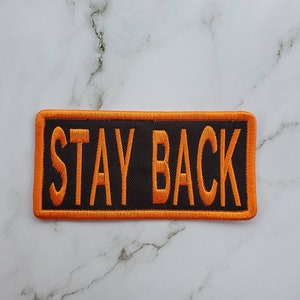 May include: Orange and black embroidered patch with the text "STAY BACK" on a black background.