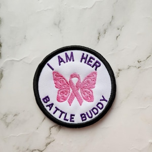 Peut inclure: Un écusson brodé blanc avec un ruban rose et un motif de papillon. Le texte "Je suis son ami de combat" est écrit en cercle autour du motif.