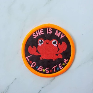 May include: A round, orange-bordered patch with a black background. The patch features a red lobster with pink eyes and a white smile. The text "SHE IS MY LOBSTER" is embroidered in pink, yellow, blue, and green around the lobster.