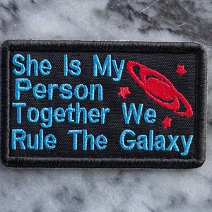 May include: Black embroidered patch with blue text that reads "She Is My Person Together We Rule The Galaxy". A red and white embroidered planet with rings is in the upper right corner.