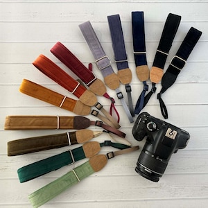 Peut inclure: Plusieurs sangles d'appareil photo colorées, rouges, oranges, marron, vertes et bleues. Chaque sangle présente une texture douce et veloutée, avec des connecteurs en liège et des éléments métalliques. Un appareil photo noir est également présent.