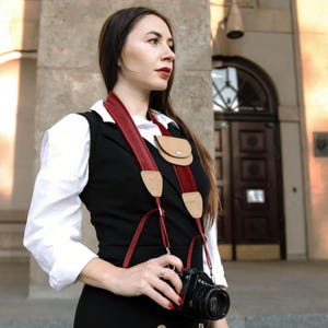 Pode incluir: Uma alça de câmera com uma faixa de couro vermelho escuro, detalhes em couro bege claro e uma pequena bolsa anexada. A alça é usada sobre uma camisa branca de colarinho e um colete preto, conectada a uma câmera fotográfica preta.