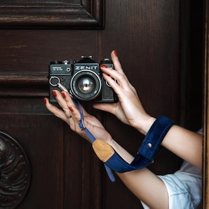 May include: A black Zenit TTL film camera with a lens attached, featuring a blue fabric camera strap with a tan leather accent piece. The strap includes a wide, padded blue velvet section and a metal adjustment buckle for securing the camera.