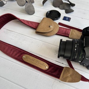 Può includere: Una tracolla per fotocamera in velluto bordeaux con dettagli in pelle marrone chiaro e una custodia abbinata. La tracolla presenta un'etichetta in pelle con "Hannah Morales Photography". Sono visibili anche un obiettivo e un tappo per fotocamera neri.