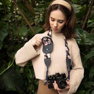 Pode incluir: Uma alça de câmara preta com um padrão floral de flores brancas, cor-de-rosa e azuis. A alça inclui uma pequena bolsa circular de cor escura presa na lateral e está ligada a uma câmara de película Zenit preta com uma lente.