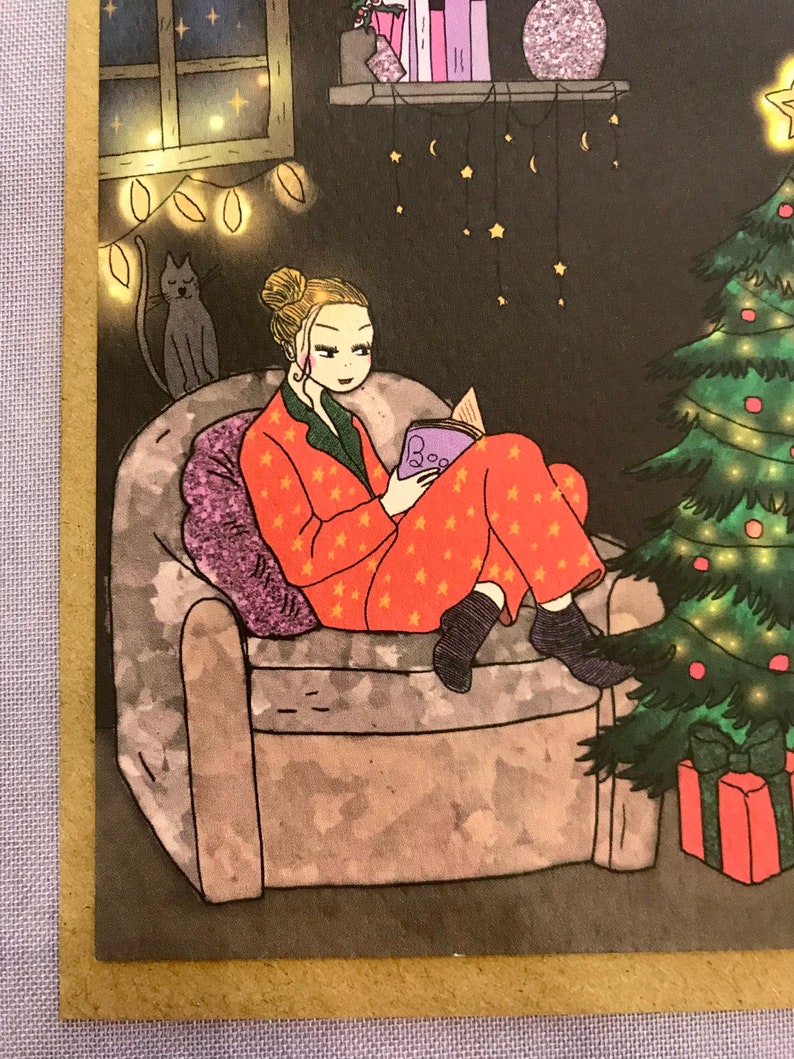 May include: A woman in red pajamas with white stars sits in a purple armchair reading a book. The book is purple with the word 'Book' on the cover. The woman is wearing black socks and black shoes. There is a cat sitting on a chair behind the woman. There is a Christmas tree with lights and ornaments in the background. There are also string lights and stars hanging from the ceiling.