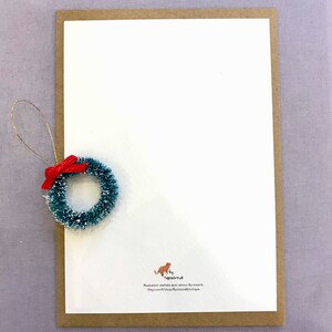 May include: A white card with a brown border. The card has a small green and red wreath ornament in the top left corner. The bottom right corner of the card has a small orange cat illustration with the text 'by agrona' and the text 'Illustration réalisée avec amour by agrona Etsy.com/shop/agronaboutique'. 