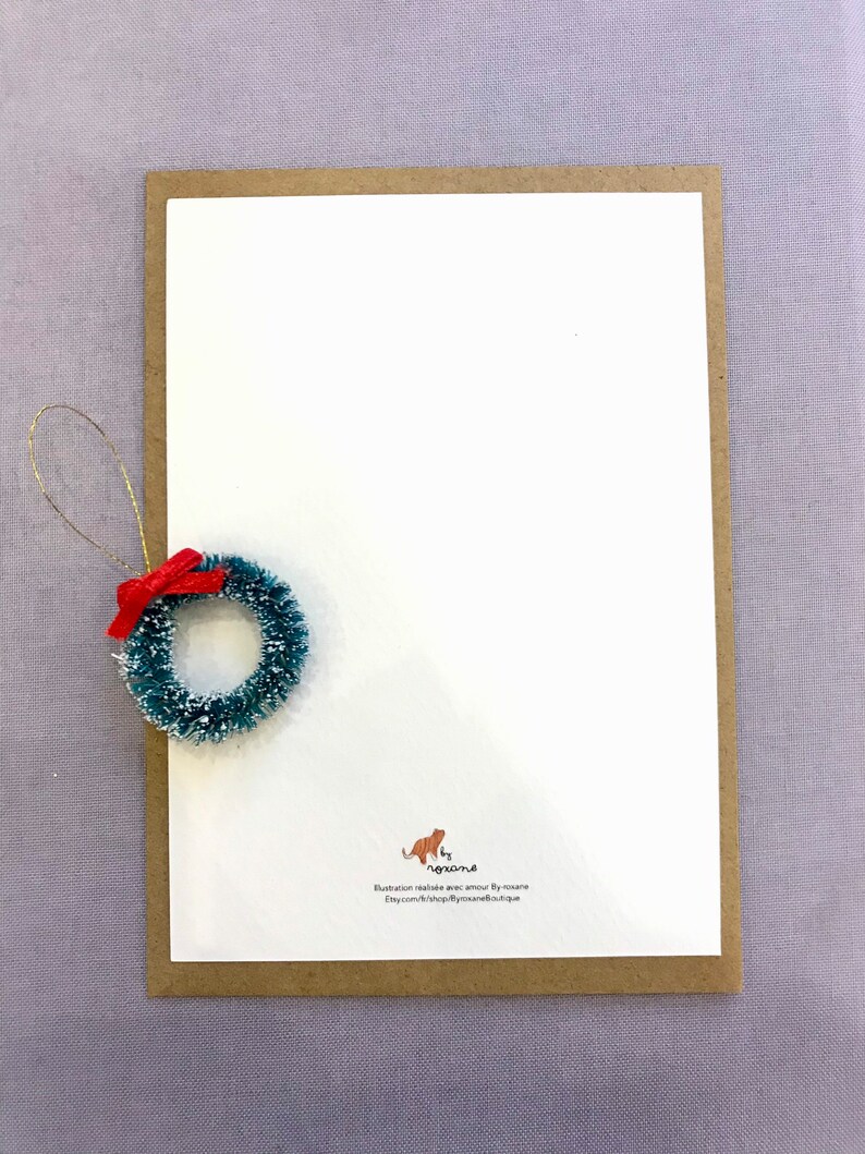 May include: A white card with a brown border. The card has a small green and red wreath ornament in the top left corner. The bottom right corner of the card has a small orange cat illustration with the text 'by agrona' and the text 'Illustration réalisée avec amour by agrona Etsy.com/shop/agronaboutique'. 