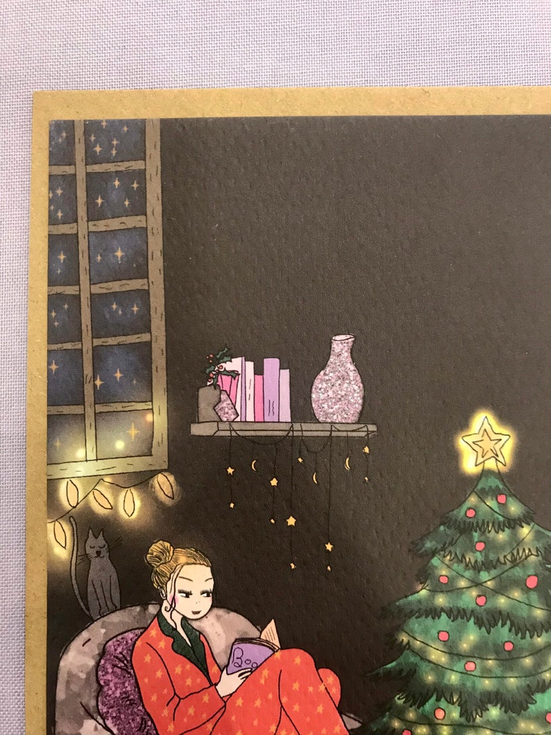 May include: A woman in red pajamas with white stars sits in a chair reading a book titled 'Boo.' The woman has blonde hair in a bun and is wearing a necklace with a small pendant. A cat sits in a chair behind the woman. A Christmas tree with green and red ornaments is in the background. The room is decorated with string lights and a shelf with a vase and books. The room is dark with a window showing a night sky with stars.