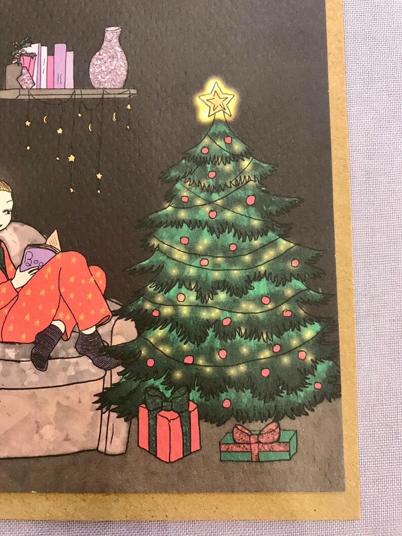 May include: A green Christmas tree with red ornaments and white lights. There are two presents under the tree, one red and one green. The tree is in a room with a person sitting in a chair reading a book.