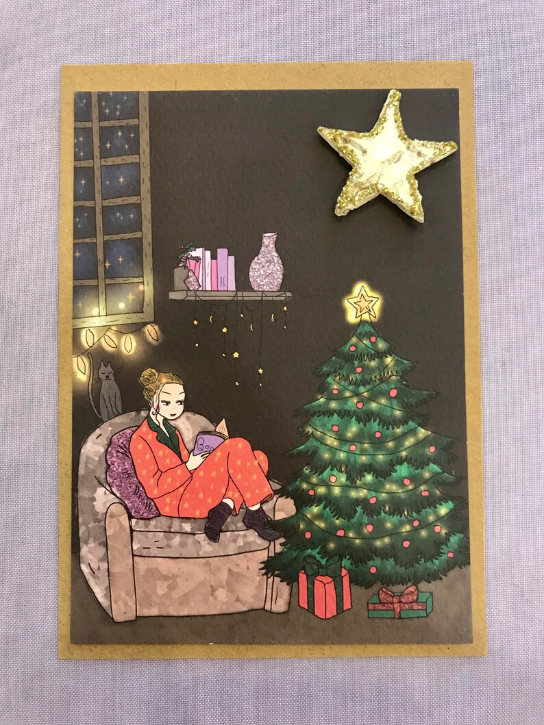 May include: A woman in red pajamas sits in an armchair reading a book. A Christmas tree with lights and ornaments is in the background. A cat sits on a chair in the background. The scene is decorated with a star, lights, and other festive decorations.