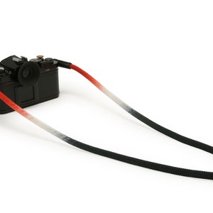 Pode incluir: Uma câmera preta com uma alça de câmera ombre vermelha, branca e preta.