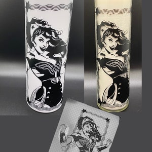 May include: A white tumbler with a black and white image of Wonder Woman. The image is framed with a black border and stars. The tumbler is next to a white candle with the same image.