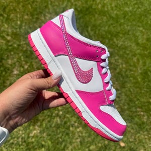 May include: A customized pink and white sneaker with a bedazzled Nike swoosh and accents. The shoe features a white sole and laces, with pink leather panels and a pink outsole. The shoe is held up against a green grassy background.