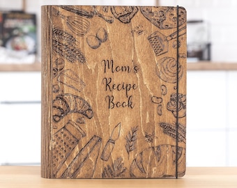 May include: A wooden recipe book with a brown wood cover and a black elastic band. The cover is decorated with a laser-engraved design of various baking and cooking ingredients, including a croissant, bread, and a whisk. The text "Mom's Recipe Book" is engraved in the center of the cover.