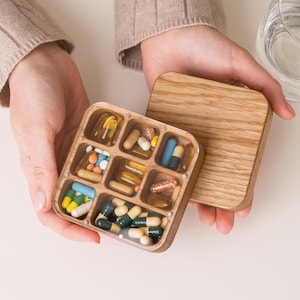 Puede incluir: Una caja de pastillas de madera con una tapa transparente, dividida en 7 compartimentos, llena de varias pastillas y cápsulas. La caja está abierta y la tapa está apoyada en la parte superior.