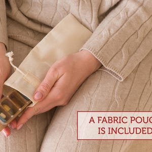 May include: A wooden pill organizer with a clear lid, filled with colorful pills. The organizer is being held by a person and is inside a white fabric pouch. The text "A FABRIC POUCH IS INCLUDED" is printed on a red rectangle below the image.
