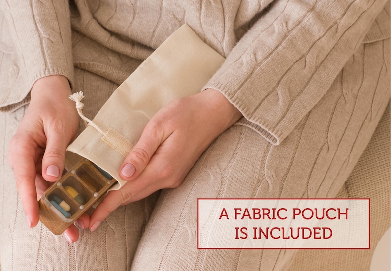 May include: A wooden pill organizer with a clear lid, filled with colorful pills. The organizer is being held by a person and is inside a white fabric pouch. The text "A FABRIC POUCH IS INCLUDED" is printed on a red rectangle below the image.