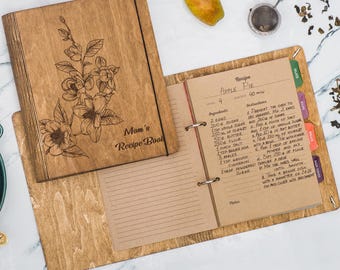 May include: A wooden recipe book with a floral design and the words "Mom's Recipe Book" engraved on the cover. The book has a ring binder and pages filled with handwritten recipes, including one for Apple Pie.
