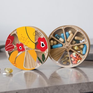 May include: A wooden pill organizer with a floral design on one side and a seven-day pill dispenser on the other side. The pill dispenser has the days of the week labeled on it.