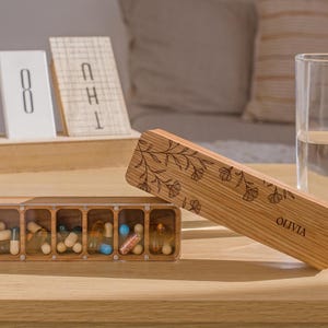 May include: A wooden pill box with seven compartments, filled with various pills. The box is engraved with a floral design and the name "Olivia".