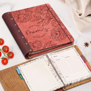 Puede incluir: Un libro de recetas de madera color coral con un lomo de cuero marrón oscuro asegurado por remaches metálicos. La cubierta presenta dibujos de líneas negras de peonías y el texto "WITH LOVE Mom's RECIPE BOOK". Una carpeta abierta muestra páginas rayadas.