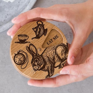 Puede incluir: Una caja redonda de madera con un conejo, una tetera, una taza y un reloj de bolsillo grabados en ella. La caja tiene las palabras "EAT ME" escritas en ella.