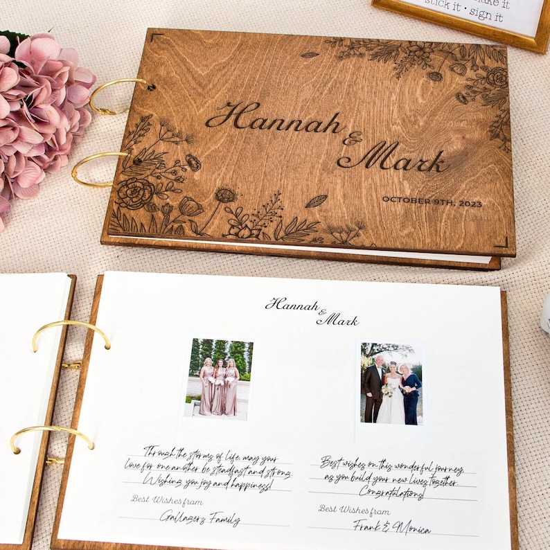 K&ouml;nnte beinhalten: Ein G&auml;stebuch aus Holz mit einem floralen Design und den eingravierten Namen "Hannah & Mark" auf dem vorderen Umschlag. Das Buch ist auf einer Seite mit zwei Fotos und handgeschriebenen Nachrichten ge&ouml;ffnet.