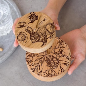 Pode incluir: Duas caixas redondas de madeira com desenhos gravados. A caixa superior apresenta um coelho, um relógio de bolso, uma xícara de chá e um bule com o texto "Eat Me". A caixa inferior apresenta um desenho floral.