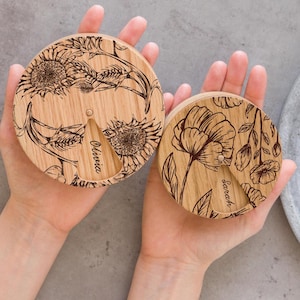 May include: Two round wooden boxes with floral designs and engraved names. The box on the left has a sunflower design and the name "Olivia" engraved on it. The box on the right has a poppy design and the name "Sarah" engraved on it.