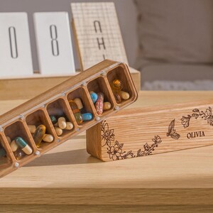 May include: A wooden pill box with seven compartments, filled with colorful pills. The box is engraved with a floral design and the name "Olivia".