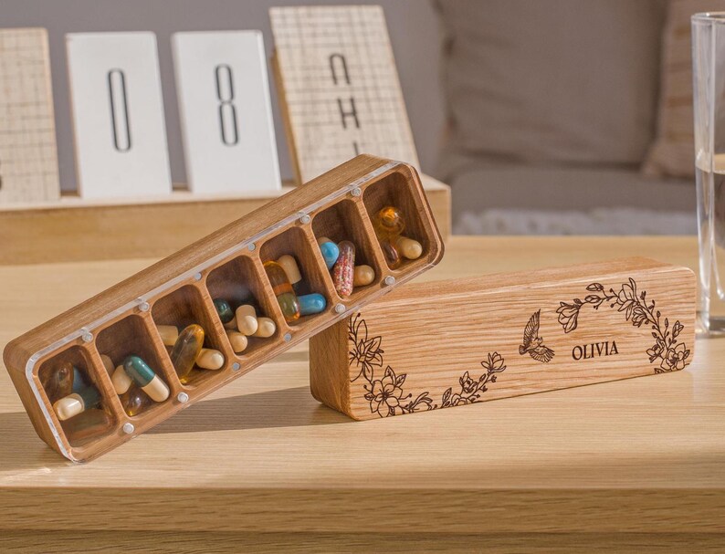 May include: A wooden pill box with seven compartments, filled with colorful pills. The box is engraved with a floral design and the name "Olivia".