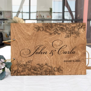K&ouml;nnte beinhalten: Ein G&auml;stebuch aus Holz mit einem floralen Design und dem Text "John & Carla" und "August 9, 2023" auf der Vorderseite.