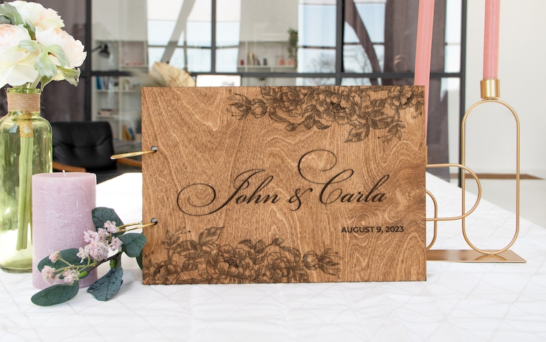 K&ouml;nnte beinhalten: Ein G&auml;stebuch aus Holz mit einem floralen Design und dem Text "John & Carla" und "August 9, 2023" auf der Vorderseite.
