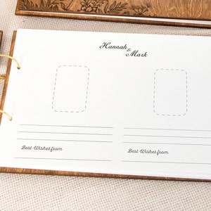 K&ouml;nnte beinhalten: Ein G&auml;stebuch aus Holz mit einem wei&szlig;en Einband und zwei Fotopl&auml;tzen. Der Einband tr&auml;gt den Text "Hannah & Mark" in Kursivschrift. Das Buch ist mit einer goldenen Ringbindung versehen und die Worte "Beste W&uuml;nsche von" sind unter den Fotopl&auml;tzen gedruckt.