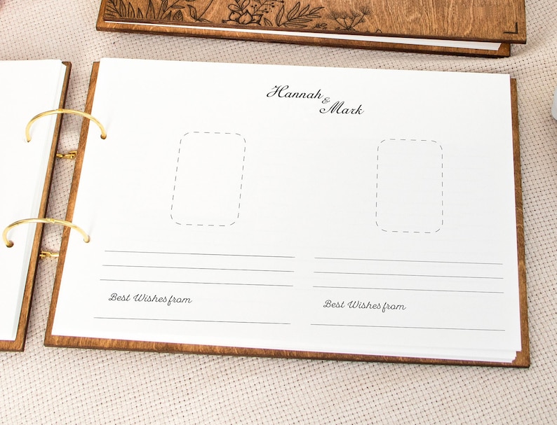 K&ouml;nnte beinhalten: Ein G&auml;stebuch aus Holz mit einem wei&szlig;en Einband und zwei Fotopl&auml;tzen. Der Einband tr&auml;gt den Text "Hannah & Mark" in Kursivschrift. Das Buch ist mit einer goldenen Ringbindung versehen und die Worte "Beste W&uuml;nsche von" sind unter den Fotopl&auml;tzen gedruckt.
