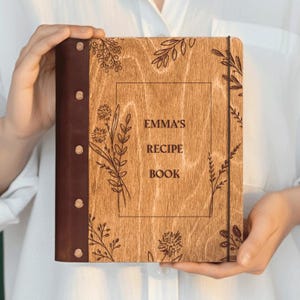 以下が含まれることがあります： 「EMMA'S RECIPE BOOK」の文字が刻まれた木製のレシピ本。表紙には花の模様があり、濃い茶色の革の背表紙に金属製のリベットが付いています。ゴムバンドで本を固定しています。