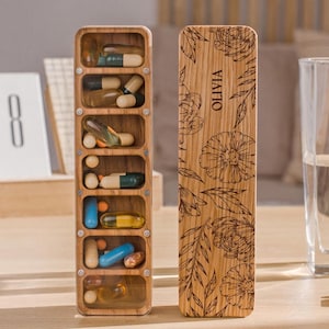 May include: A wooden pill organizer with seven compartments, each containing different colored pills. The organizer is personalized with the name "Olivia" and a floral design.