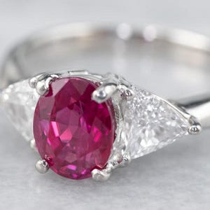 May include: A silver ring with a central oval ruby and two flanking triangular clear gemstones. The ruby is a deep red colour. The ring's band is smooth and polished. The gemstones are set in a silver metal setting, reflecting light. The ring is on a light background.