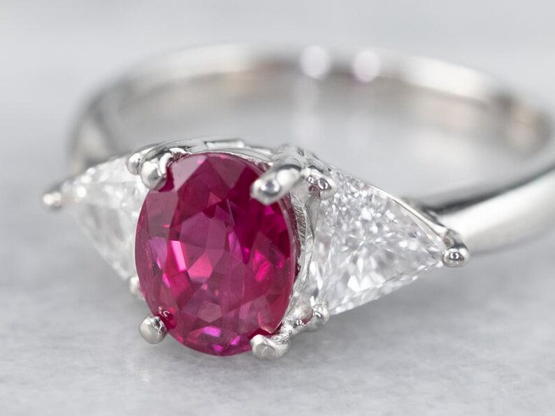 May include: A silver ring with a central oval ruby and two flanking triangular clear gemstones. The ruby is a deep red colour. The ring's band is smooth and polished. The gemstones are set in a silver metal setting, reflecting light. The ring is on a light background.