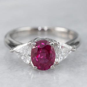May include: A silver ring with a large, oval-cut, deep red gemstone and two triangular, clear gemstones. The ring's band is smooth and polished, and the setting is a bright silver colour.