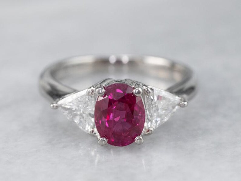 May include: A silver ring with a large, oval-cut, deep red gemstone and two triangular, clear gemstones. The ring's band is smooth and polished, and the setting is a bright silver colour.