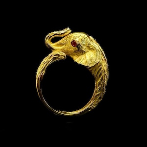 Puede incluir: Un anillo dorado con forma de elefante, con un ojo de piedra roja. La trompa del elefante se curva para encontrarse con la banda del anillo. El anillo está sobre un fondo negro.
