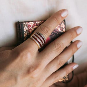 Op de afbeelding: Een goudkleurige ring met een gebogen ontwerp, met afwisselende rijen rode edelstenen en heldere kristallen. De ring wordt aan een vinger gedragen en toont zijn ingewikkelde details en levendige kleuren. Het ontwerp van de ring is opvallend en elegant.